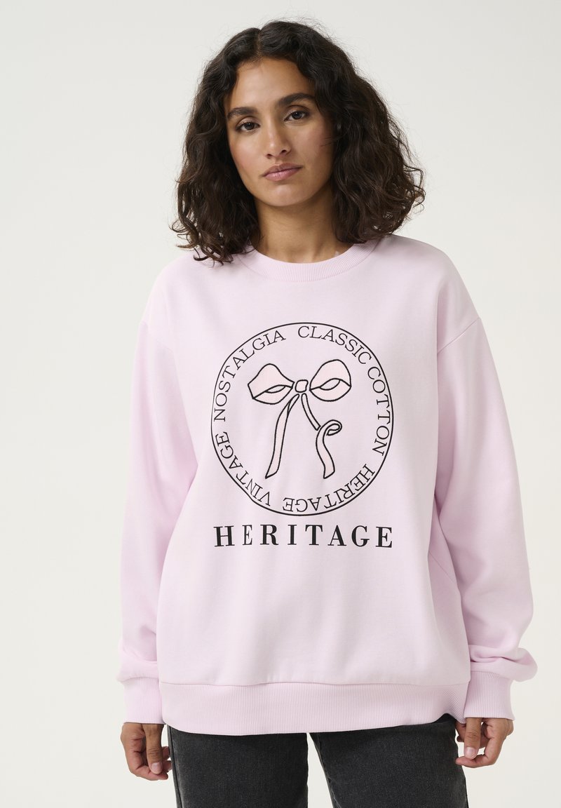Maglione rosa chiaro in cotone, con un design grafico circolare che presenta un fiocco e la scritta "HERITAGE" in nero. Tessuto morbido, vestibilità rilassata.