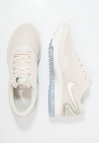 Une paire de chaussures de course Nike beiges avec une texture en maille, des lacets et une semelle à coussin d'air visible sur fond blanc.