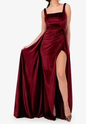 Kvinde iført ærmeløs mørkerød fløjlskjole med firkantet udskæring og høj slids, der holder kjolens side, kombineret med bronzefarvede spidse hæle.