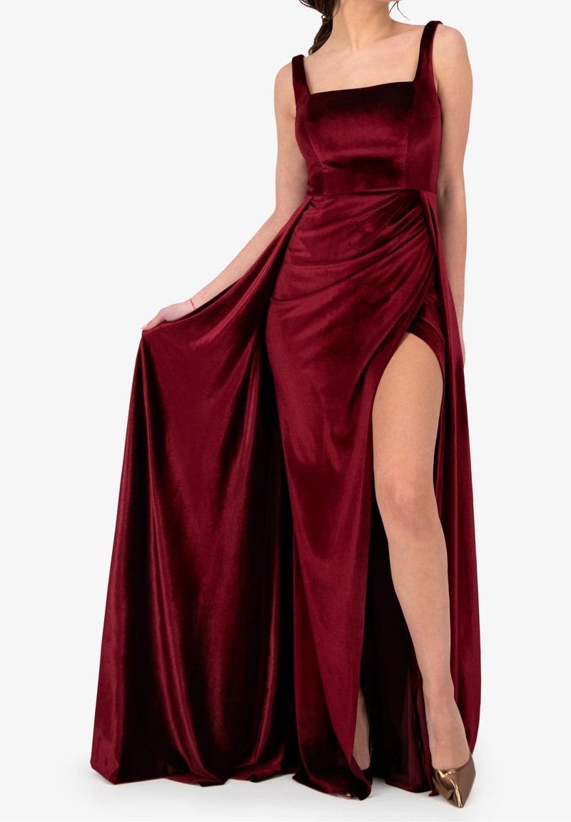 Femme portant une robe sans manches en velours rouge foncé avec décolleté carré et fente haute, tenant le côté de la robe, assortie à des talons pointus bronze.