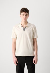 Cremfarbener Polo-Shirt mit kurzen Ärmeln, versehen mit einem schwarz-weiß gestreiften Kragen und einem kleinen Logo auf der linken Brust.