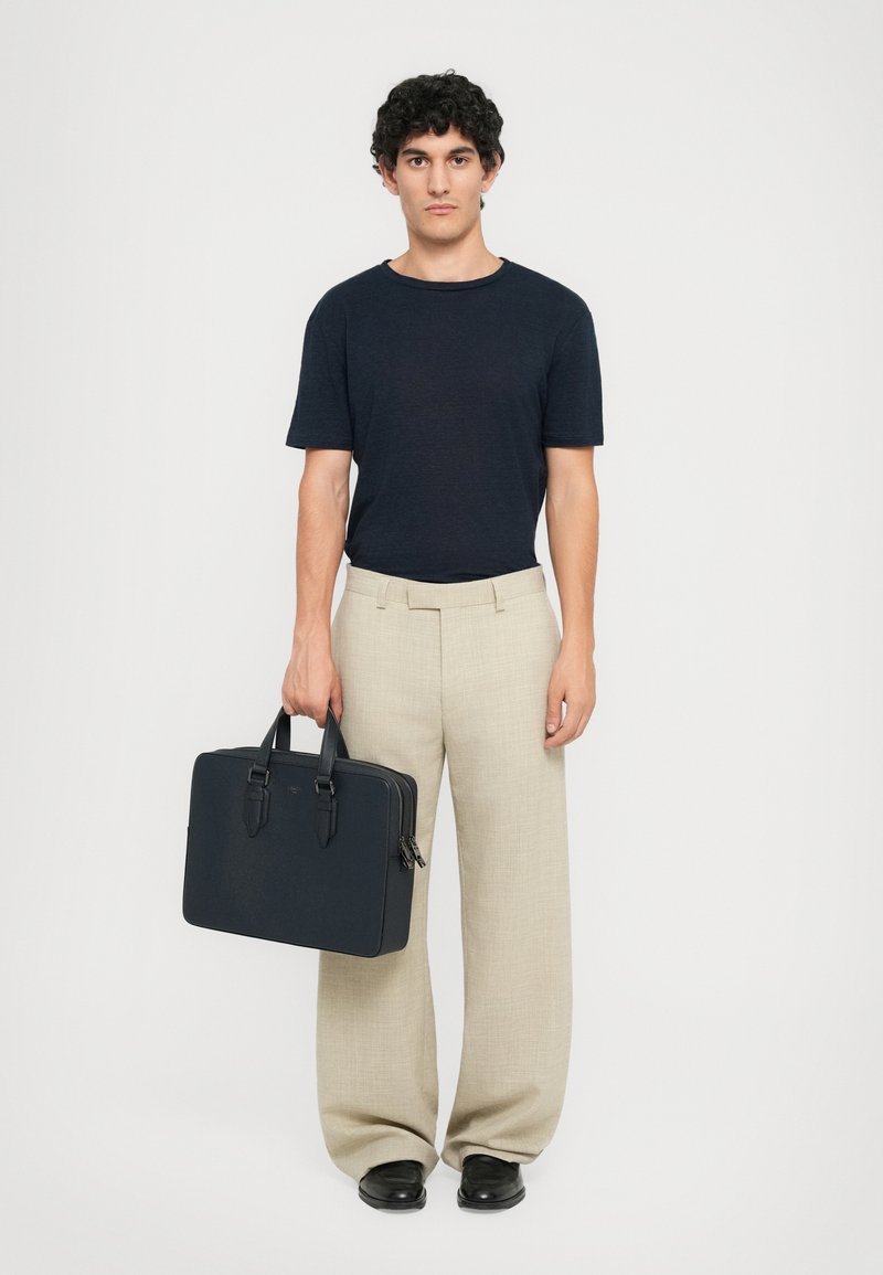 Chemise à manches courtes bleu foncé, pantalon large beige et sac pour ordinateur portable texturé noir avec double poignées. Des chaussures noires complètent le look.