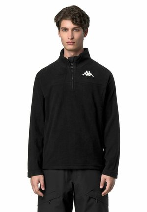 Giovane uomo che indossa un pile nero con cerniera a quarto e logo bianco sul petto, pantaloni cargo neri, in piedi davanti a uno sfondo bianco.