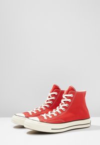 Red high-top sneakers made of canvas, featuring white laces, rubber toe caps, and contrasting black stripes on the sole.