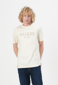 Pamučna majica boje kreme s logotipom "GUESS EST. 1981" u bež boji. Kratki rukavi, okrugli izrez i ravni rub. Nosena s tamnim trapericama.