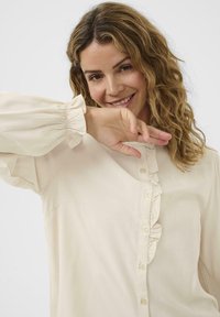 Femme avec des cheveux blonds ondulés souriante, portant un blouse beige avec une patte de boutonnage froncée et de longues manches ballons sur fond blanc.