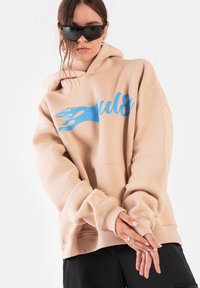 Beżowa, oversize bluza z kapturem z niebieskim grafika płomieni, z prążkowanymi mankietami i dołem. Model w czarnych okularach przeciwsłonecznych, z rękami skrzyżowanymi na przodzie.