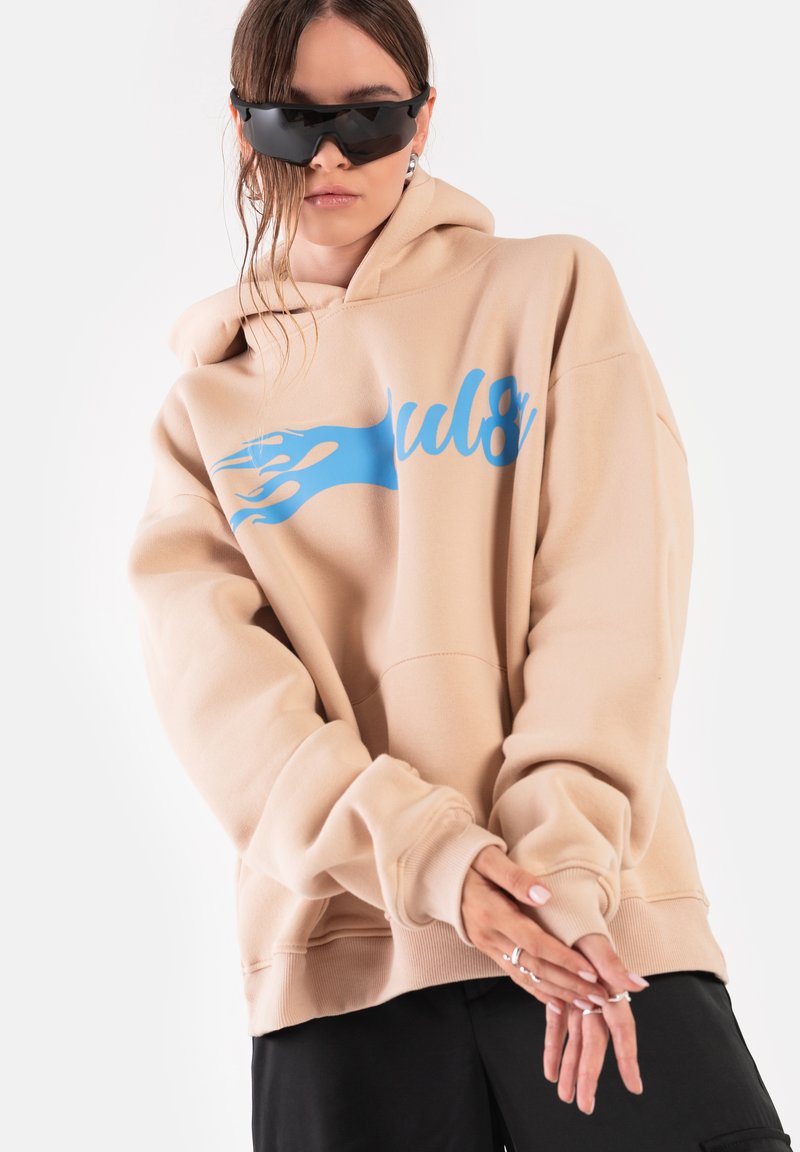 Beżowa, oversize bluza z kapturem z niebieskim grafika płomieni, z prążkowanymi mankietami i dołem. Model w czarnych okularach przeciwsłonecznych, z rękami skrzyżowanymi na przodzie.