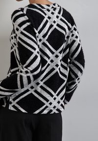 Osoba ubrana w sweter w czarno-białe geometryczne wzory z długimi rękawami, pokazana od tyłu od pasa w górę na szarym tle.