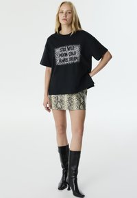 Camiseta negra de manga corta con texto en blanco, combinada con una minifalda de estampado de serpiente metálico y botas de charol negras hasta la rodilla.