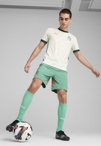 Equipación de fútbol Puma que incluye una camiseta blanca con patrones claros, pantalones cortos verdes y calcetines a juego; botas negras con un balón estilizado.