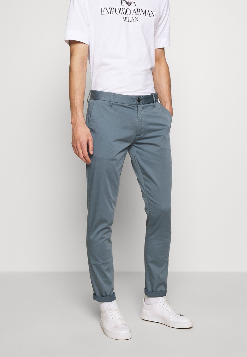 Homme portant un pantalon slim bleu clair, un t-shirt blanc Emporio Armani, des baskets blanches, debout avec une main dans la poche.