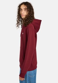 Sweat à capuche bordeaux avec cordon de serrage, poche avant, poignets et ourlet côtelés, fabriqué en tissu doux et texturé. Présenté de côté.