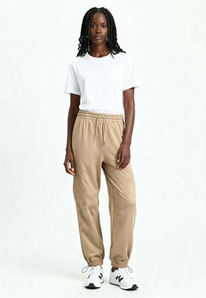 Femme avec de longues tresses portant un t-shirt blanc, un pantalon de jogging beige et des baskets blanches, debout devant un fond blanc uni.