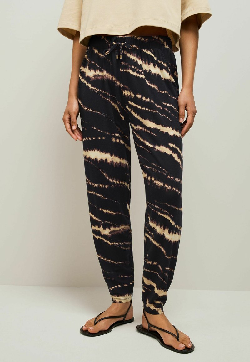 Zwarte tie-dye joggers met beige strepen, voorzien van een elastische tailleband, zijzakken en een relaxte fit, gecombineerd met zwarte flip-flops.