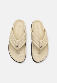 Beige lærflip-flops med et krydset design, der har en lille dekorativ metalaccent og en sort gummisål.