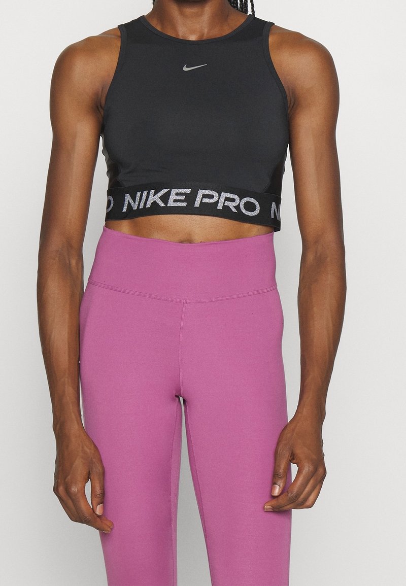 Femme portant un crop top Nike Pro noir et un legging violet taille haute, debout devant un fond blanc.