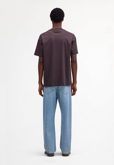 Mann steht mit dem Rücken zur Kamera, trägt ein dunkelbraunes kurzärmeliges T-Shirt, hellblaue Jeans mit weitem Bein und dunkle Schuhe vor einem weißen Hintergrund.