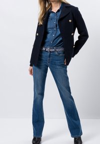 Blazer croisé en laine mélangée marine avec des boutons dorés, associé à une chemise en denim et un jean évasé bleu, accessoirisé d'une ceinture à motifs.