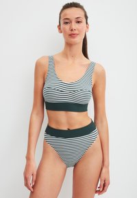 Vrouw die tegen een witte achtergrond staat, gekleed in een groen-wit gestreepte sportbeha en hoge tailleslip, kijkend naar de camera met een neutrale uitdrukking.