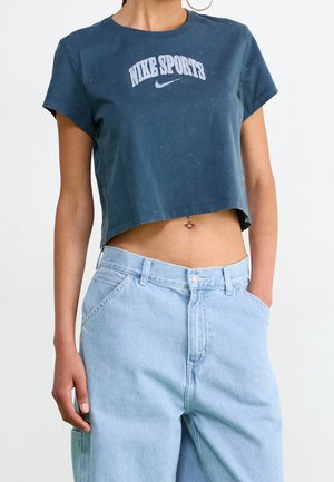 Kobieta nosząca krótką granatową sportową koszulkę Nike, jasnoniebieskie jeansy z wysokim stanem i widoczny kolczyk w pępku.
