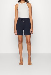 Pantalones cortos de algodón azul marino con una cinturilla elástica y cordón, combinados con una blusa sin mangas de color gris claro. Sandalias de tacón cuadrado en tono marrón claro.