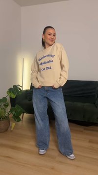 Felpa beige con scritta blu "Timberland Authentic Est 1973", abbinata a jeans a gamba larga azzurri e sneakers grigie. Ambientazione interna.