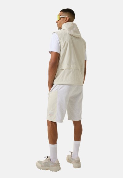 Beige ärmellose Jacke mit Kapuze, weißes T-Shirt, graue Shorts, beige Sneakers und gelbe Sonnenbrille. Mit einer Seitentasche und Logodetail.