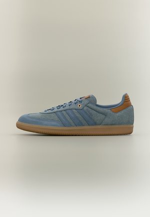 Zapatilla deportiva azul casual con lengüeta marrón en el talón y suela de goma, vista lateral, mostrando tejido texturizado y diseño con cordones sobre fondo liso.