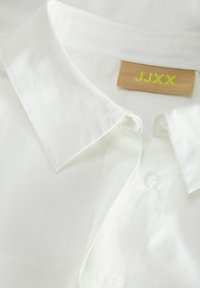Witte button-up shirt met een gestructureerde kraag, voorzien van een beige label met gele tekst en doorzichtige knopen. Glad, lichtgewicht stoftextuur.