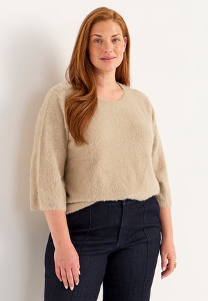 Vaalean beige pörröinen neulepaita, jossa on pyöreä pääntie ja kolmannes hihat, yhdistettynä tummiin farkkuihin. Pehmeä rakenne, yksinkertainen muotoilu.