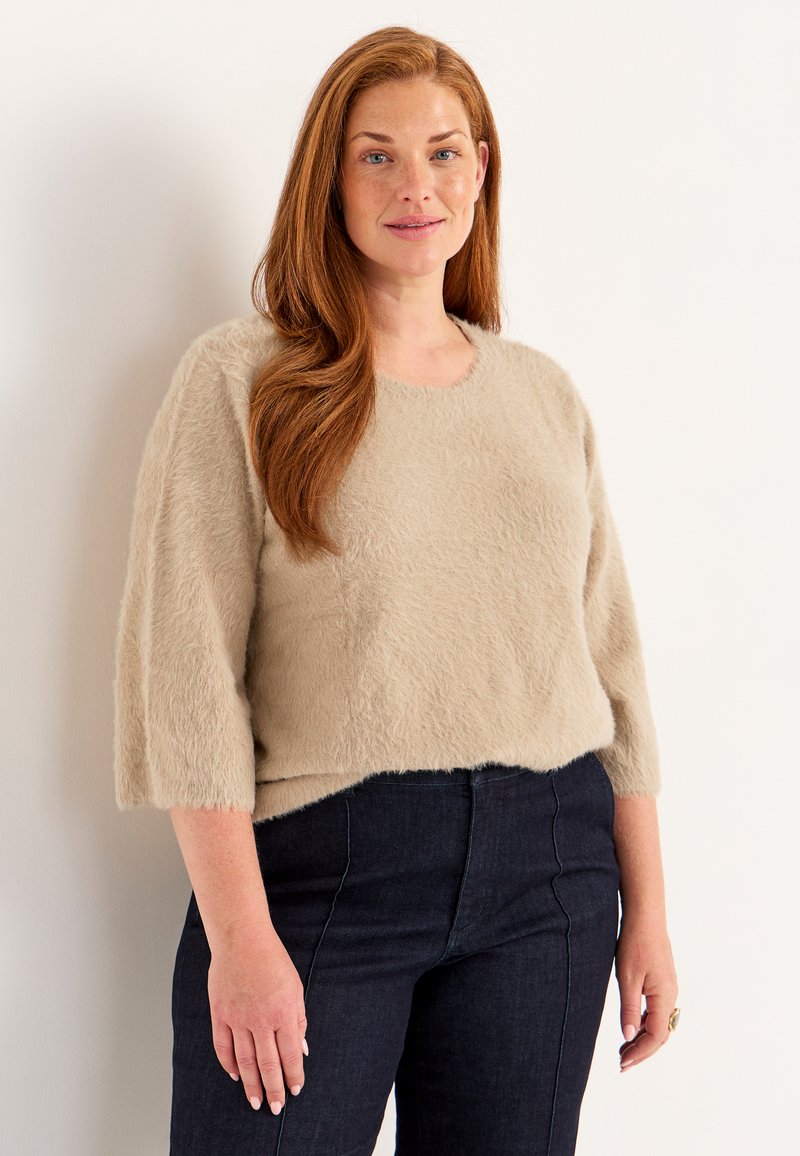 Beige, flauschiger Strickpullover mit rundem Ausschnitt und drei Viertelärmeln, kombiniert mit dunkelblauen Jeans. Weiche Textur, einfaches Design.