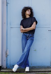 Schwarzes kurz geschnittenes T-Shirt, blaue weit geschnittene Jeans und weiße Sneakers. Modell mit lockigem Haar lehnt an einer hellblauen Tür. Einfache Accessoires.