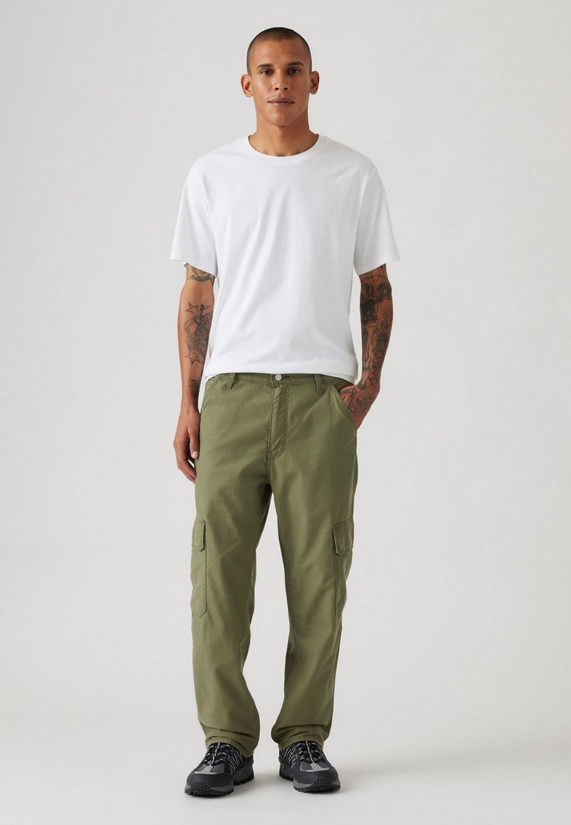 T-shirt bianca in cotone e pantaloni cargo verde oliva con tasche laterali. Scarpe sportive nere. Design semplice, vestibilità comoda, texture liscia.