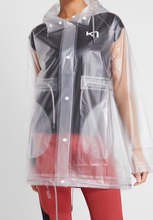 Person trägt einen transparenten Regenmantel mit weißen Knöpfen und Kordelzügen über einem schwarzen Oberteil und roten Hosen vor einem einfarbigen Hintergrund.