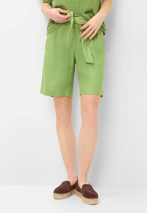 Person iført grønne shorts med snøre i taljen, matchende grøn top og brune espadrillos slip-on sko, stående mod en ensfarvet hvid baggrund.