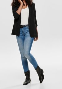 Blazer nero sopra una maglietta bianca, abbinato a jeans skinny azzurri chiari e stivali alla caviglia neri. Tessuto morbido e design strutturato con dettagli sottili.