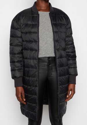 Person trägt eine lange schwarze gesteppte Jacke über einem grauen Hemd und hoch taillierte schwarze Hosen mit Knopfdetails, steht vor einem schlichten Hintergrund.