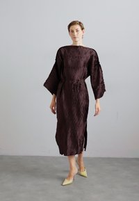 Brown, textured, long-sleeve dress with a loose fit and waist tie. Features a round neckline and side slits. Worn with light yellow heels.