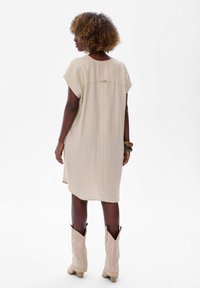 Vrouw met krullend haar, gekleed in een beige jurk tot op de knie, lichte beige laarzen en armbanden, staande tegen een effen witte achtergrond.