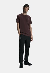 T-shirt bordeaux à manches courtes avec un logo sur la poitrine, associé à un pantalon noir avec des poches zippées et des chaussures noires à semelles texturées.