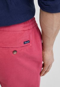Pantalon rose à cordon de serrage avec une texture uniforme, doté d'une poche arrière avec un bouton et d'une petite étiquette logo. Le tissu semble doux et léger.