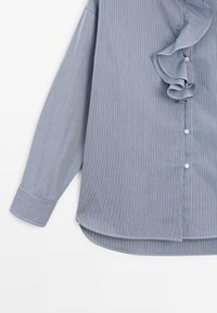 Světle modrá a bílá proužkovaná košile s dlouhým rukávem, dekorací s volánky vpředu a bílými knoflíky, manžety na rukávech a zaobleným lemem.