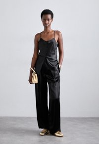 Black satin camisole with buttons, paired with loose black satin pants. Model carries a small cream bag and wears gold shoes. Smooth texture.