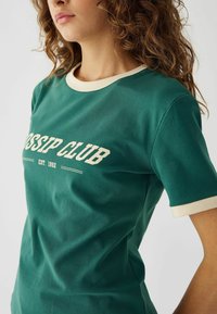 Zelena majica s kratkimi rokavi in kremastim robom, ki nosi napis "GOSSIP CLUB EST. 1992" v belem tisku. Mehak bombažen material.