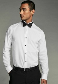 Cămașă albă cu nasturi negri, mâneci lungi și un papion negru. Prezintă un guler bine conturat și o textură netedă, purtată cu pantaloni negri.