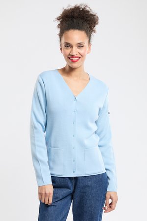 Femme souriante aux cheveux bouclés portant un cardigan bleu clair boutonné avec des poches et un jean bleu, debout devant un fond uni.