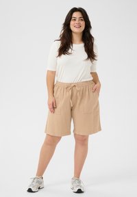 Beige cotton shorts featuring an elastic waistband with a drawstring, two side pockets, and a relaxed fit, paired with a white long-sleeve top.
