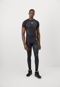 Sort atletisk kortærmet top og leggings med guld accenter og mønstre, der har et tætsiddende design og glat tekstur. Sorte sneakers.
