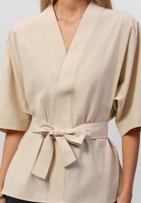 Top in stile kimono beige realizzato in tessuto leggero, con maniche larghe e cintura da annodare in vita, caratterizzato da una sottile trama e design a scollo a V.